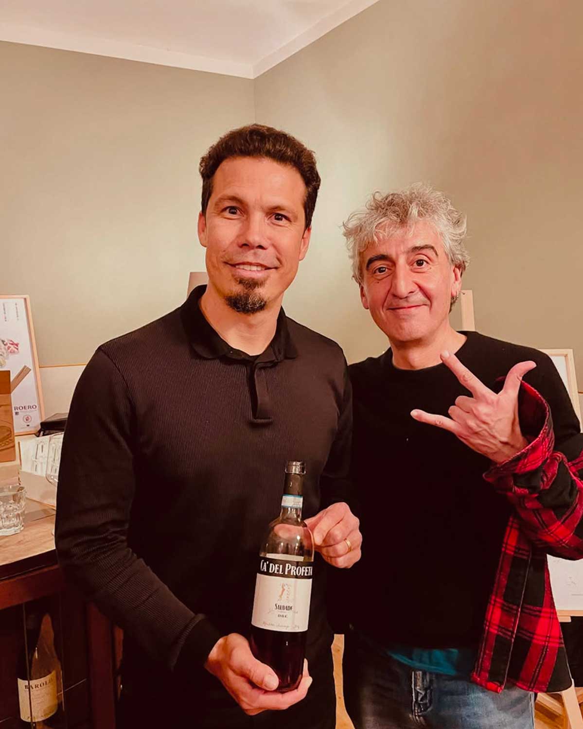 Due uomini sorridono, uno con barba tiene una bottiglia di vino rosso "Saudade Ca' del Profeti", l'altro con capelli grigi fa un gesto rock. Sfondo interno.