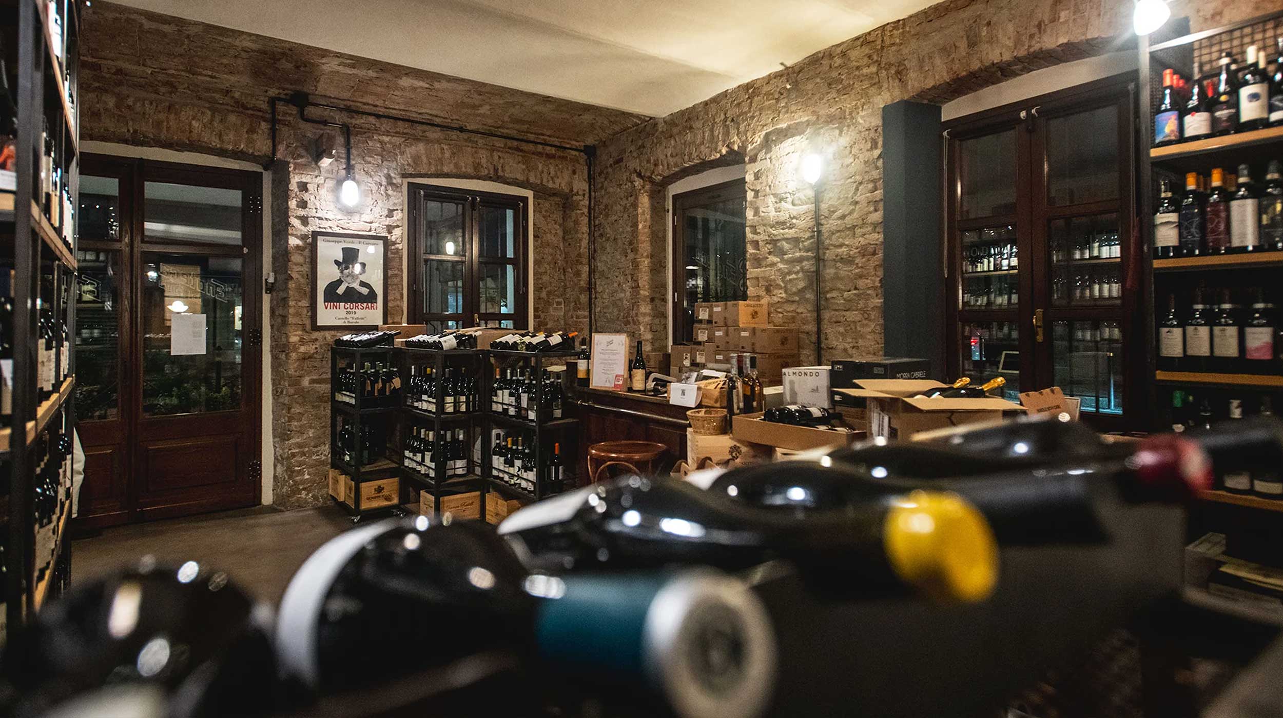 Interno di un'enoteca con pareti in mattoni a vista, scaffali e casse di vino. Bottiglie in primo piano sfocate. Poster "VINI CORSARI".
