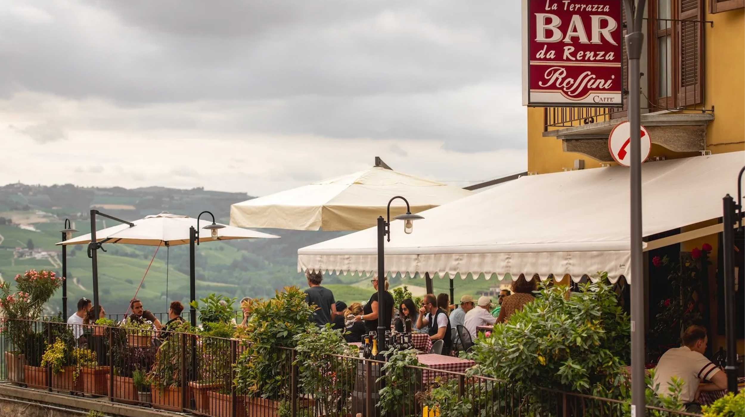 Terrazza di un bar-ristorante con persone che pranzano sotto ombrelloni, affacciata su colline verdi. Insegna "BAR da Renzo Roffini". Cielo nuvoloso.